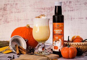 Auf dem Foto ist eine Flasche Pumpkin Spice Sirup und ein Pumpkin Spice Latte zu sehen. Im Hintergrund liegt ein Kürbis und viele Gewürze. | © SONNENTOR