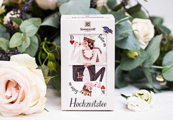 Foto von einer Packung Hochzeitstee. Der Tee steht auf einem Tisch mit Blumen im Hintergrund. | © SONNENTOR