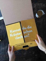 Aufnahme eines halb geöffneten, sonnengelben SONNENTOR Versandkartons mit Unterschrift der Person, die den Karton gepackt hat. | © SONNENTOR