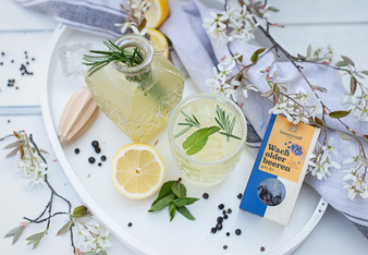 Das Foto zeigt eine Kristall- Flasche mit verdünntem Tschin Tschin Sirup, nebenbei ein Glas. Beides steht auf einem Tablett, umgeben von einem Blütenzweig, einer Zitronenscheibe und einer Packung Wacholderbeeren. | © SONNENTOR