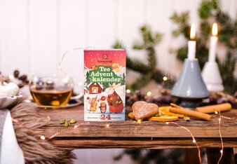 Der Teeadventkalender mit den Weihnachtstieren drauf steht auf einem weihnachtlich dekorierten Tisch Mit Kerzen, einer Lichterkette, Zimtstangen und Orangenscheiben.  | © SONNENTOR