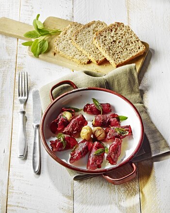 Auf dem Foto ist eine Pfanne mit Maroni-Saltimbocca zu sehen. Daneben sieht man auf einem Holzbrett drei Scheiben Brot. | © SONNENTOR