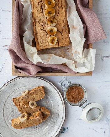 Auf dem Foto ist ein Bananen Zimt Brot mit Bananen garniert zu sehen. Davor liegen auf einem Teller aufgeschnittene Brotscheiben. | © SONNENTOR