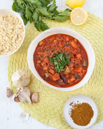 Auf dem Foto ist ein Teller mit Kichererbsen-Gemüse-Tajine zu sehen. Daneben sieht man Knoblauch, Gewürze, Zitronen und Petersilie. | © SONNENTOR