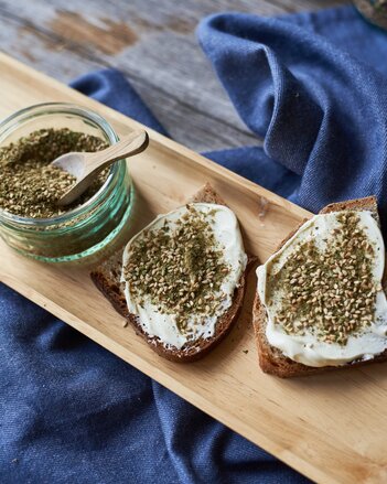 Auf dem Foto sieht man ein Gläschen mit Zatar. Daneben liegen zwei Scheiben Brot mit Aufstrich und Zatar darauf. | © SONNENTOR