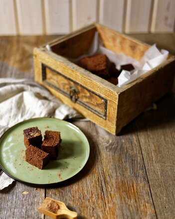 Auf dem Foto sind drei Stück Brownies auf einem grünen Teller zu sehen. Dahinter steht ein Kistchen mit mehreren Brownies darin. | © SONNENTOR