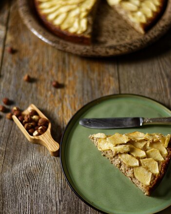 Auf dem Foto ist ein Stück Apfelkuchen zu sehen. Daneben sieht man Haselnüsse und im Hintergrund steht der restliche Kuchen. | © SONNENTOR