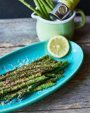 Auf dem Foto ist ein blauer Teller mit grünem Spargel darauf zu sehen. Der Spargel ist mit Parmesan bestreut, und daneben liegt eine Zitronenscheibe. | © SONNENTOR