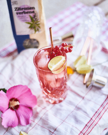 Auf dem Foto sieht man ein Glas mit Vodka-Preiselbeer. Im Hintergrund sieht man eine Packung SONNENTOR Zitronenverbene Tee. | © SONNENTOR