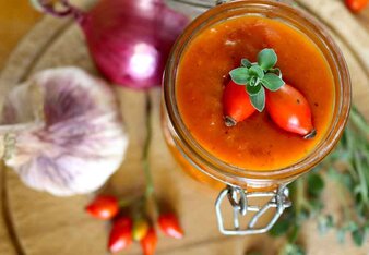 Auf dem Foto sieht man ein Glas Hagebutten Ketchup auf einem Holzbrett stehen. Auf dem Brett sieht man auch Zwiebeln, Hagebutten und Kräuter. | © SONNENTOR