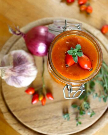 Auf dem Foto sieht man ein Glas Hagebutten Ketchup auf einem Holzbrett stehen. Auf dem Brett sieht man auch Zwiebeln, Hagebutten und Kräuter. | © SONNENTOR