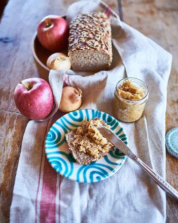 Auf dem Foto ist zu sehen, wie de Apfel-Zwiebel-Aufstrich auf ein Brot gestrichen wird. Daneben ist das Brot und das Glas mit Aufstrich zu sehen. | © SONNENTOR