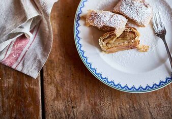 Auf dem Foto ist ein Teller mit Apfelstrudel zu sehen. Er ist mit Staubzucker bestäubt. Dahinter steht ein schwarzes Bleich mit zwei Apfelstrudel.  | © SONNENTOR