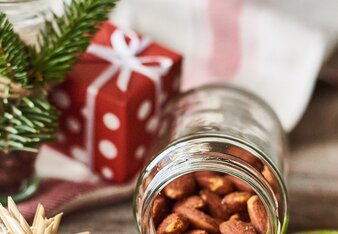 Auf dem Foto sieht man ein Glas mit süßen Gewürzmandeln. Daneben sind viele Weihnachtliche Gegenstände, wie Holzsterne, zu sehen. | © SONNENTOR