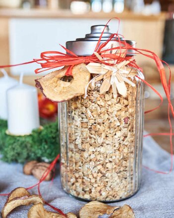 Foto von einem Glas Apfelstrudel-Granola. Das Glas ist mit roten Bändern dekoriert. Davor sind getrocknete Apfelringe zu sehen. | © SONNENTOR