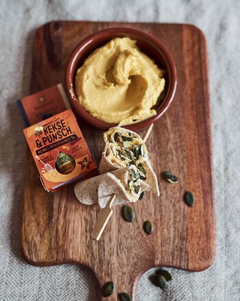 Auf dem Foto ist ein Holzbrett mit einer Schale Kürbis-Hummus darauf zu sehen. Davor liegen Mini-Wraps. | © SONNENTOR