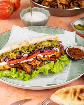 Der Tofu Kebab ist von oben geschichtet gefüllt mit grünem Paprika, roten Zwiebeln, Tomatenscheiben, Sauce, Tofu und einem Salatblatt und liegt auf einem Teller neben einem Schüsselchen Harissa-Gewürz. Im Hintergrund steht eine weiße Dip-Sauce in einem Schälchen und man sieht 2 Paradeiser.  | © SONNENTOR