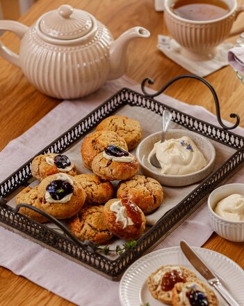 Die Scones liegen auf einem Tablett mit schwarzem Rand und schwarzen Hänkeln, ein Schälchen Clotted Cream steht ebenso darauf. Manche Scones sind mit Clotted Cream und Fruchtaufstrich dekoriert. Neben dem Tablett steht eine Teekanne und eine Tasse Tee. | © SONNENTOR