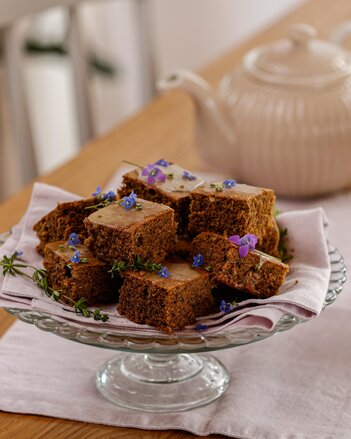 Die Schwarzteekuchen Stückchen liegen auf einer Etagere und sind mit kleinen blauen und violetten Blümchen dekoriert. Im Hintergrund steht eine Teekanne.  | © SONNENTOR