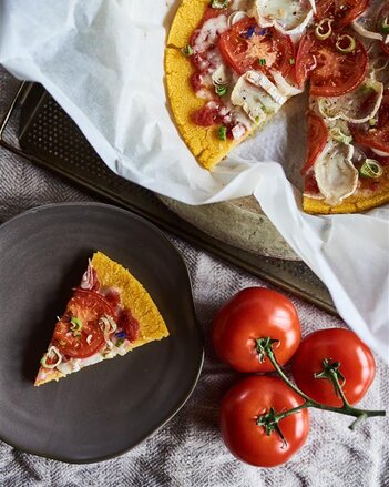 Auf dem Foto sieht man eine Polenta Pizza. Ein Stück wurde heraus genommen und liegt auf einem schwarzen Teller. Daneben sind Tomaten zu sehen. | © SONNENTOR