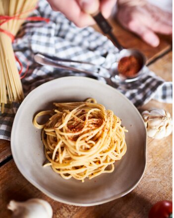 Auf dem Foto ist ein Teller mit Spaghetti aglio e olio zu sehen. Neben dem Teller sieht man Knoblauch, Tomaten und ungekochte Nudeln. | © SONNENTOR