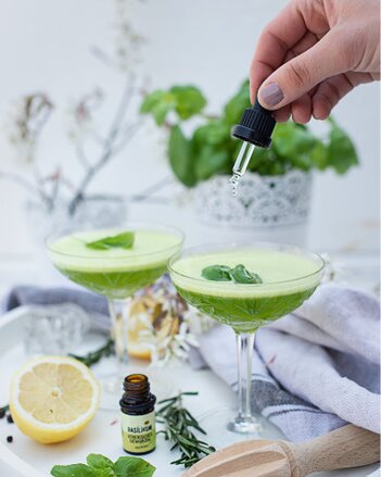 Auf dem Foto sind zwei Cocktailgläser mit grüner Basilikum Limonade zu sehen. Man sieht wie ein Gewürzöl in das Getränk getropft wird. | © SONNENTOR