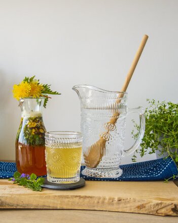 Auf dem Foto ist eine Karaffe mit Frühlingskräuter Oxymel zu sehen. Darin sind zahlreiche Kräuter. Nebenbei steht ein Krug Wasser. | © SONNENTOR