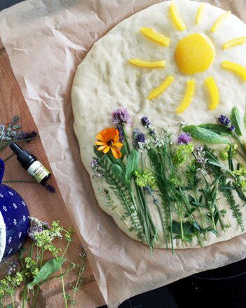 Auf dem Foto ist eine Focaccia zu sehen, auf der verschiedene Kräuter zu sehen sind. Im oberen Teil ist eine Sonne dargestellt. | © SONNENTOR