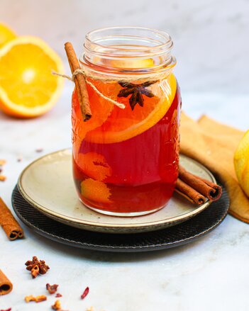 Der Orangenpunsch ist in ein Schraubglas gefüllt, das auf zwei Tellern steht, darin schwimmen Orangenschalen, eine Orangenscheibe und ein Sternanis. Dekoriert ist das Glas mit ganzen Zimtstangen. | © SONNENTOR