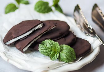 Vier Pfefferminzblättchen sind mit dunkler Schokolade überzogene Taler und liegen auf einem weißen Teller, eines ist auseinandergebrochen und man sieht die weiße Füllung. Ein Pfefferminzblatt liegt zur Garnitur daneben.  | © SONNENTOR