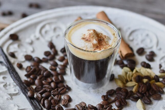 Auf dem Foto sieh man ein Glas mit einem Espresso macchiato darin. Daneben liegen Kaffeebohnen, Zimtstangen und Vanilleschoten. | © SONNENTOR